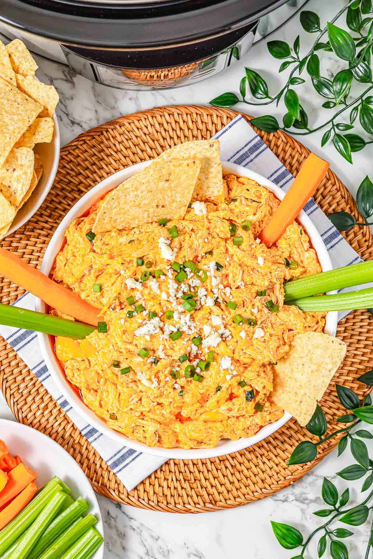 A bowl of creamy Buffalo Chicken Dip made in the crockpot, topped with chives and cheese, surrounded by celery, carrots, and tortilla chips.