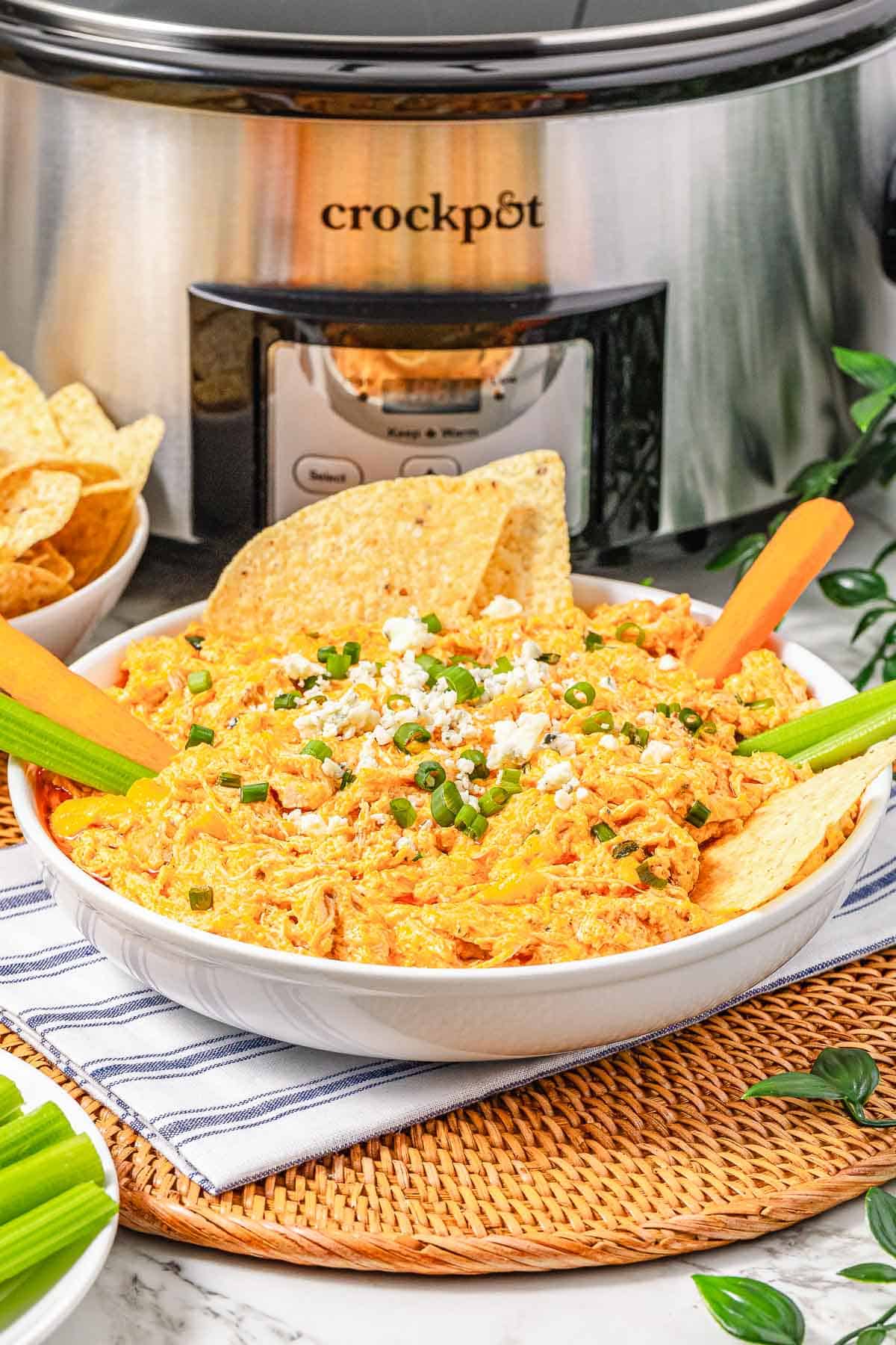 A bowl of creamy Buffalo Chicken Dip topped with green onions and cheese sits next to chips and celery, all in front of a Crockpot.