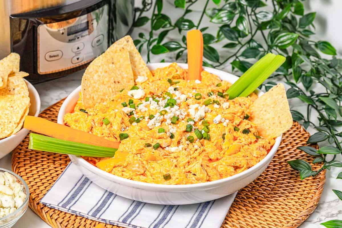 A bowl of creamy Buffalo Chicken Dip topped with green onions, served alongside celery, carrot sticks, and crunchy tortilla chips.