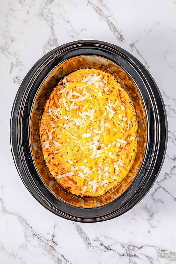 Shredded cheese in a Crockpot on a white marble countertop, ready to be cooked for delicious Buffalo Chicken Dip.