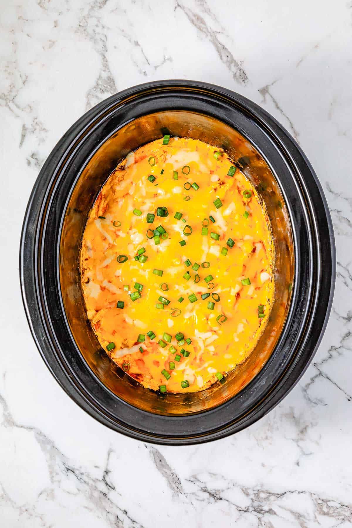 Cheesy Buffalo Chicken Dip casserole topped with chopped green onions, served in a black Crockpot on a white marble counter.