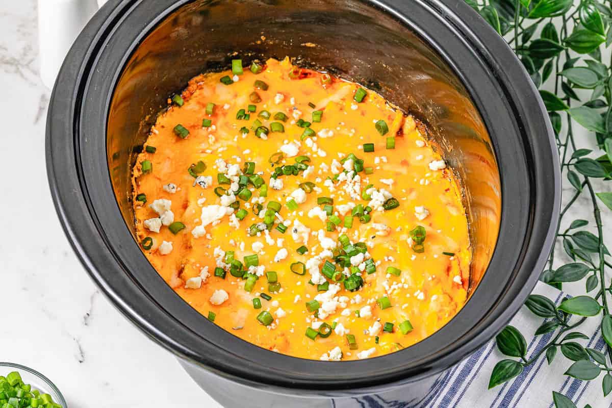 Crockpot Buffalo Chicken Dip topped with green onions and crumbled cheese in a black slow cooker on a marble countertop.