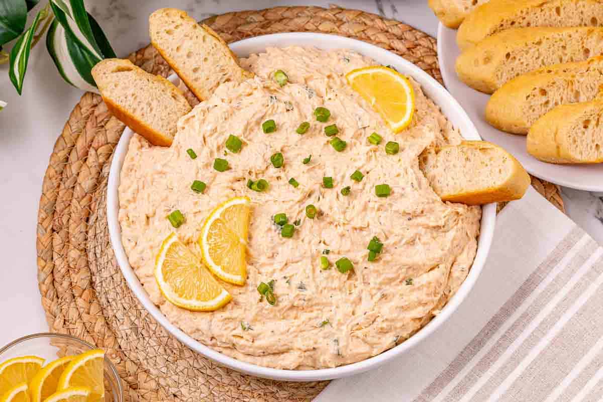 Bowl of creamy crab dip garnished with lemon slices, green onions, and bread slices, on a woven placemat&mdash;an easy recipe perfect for entertaining.