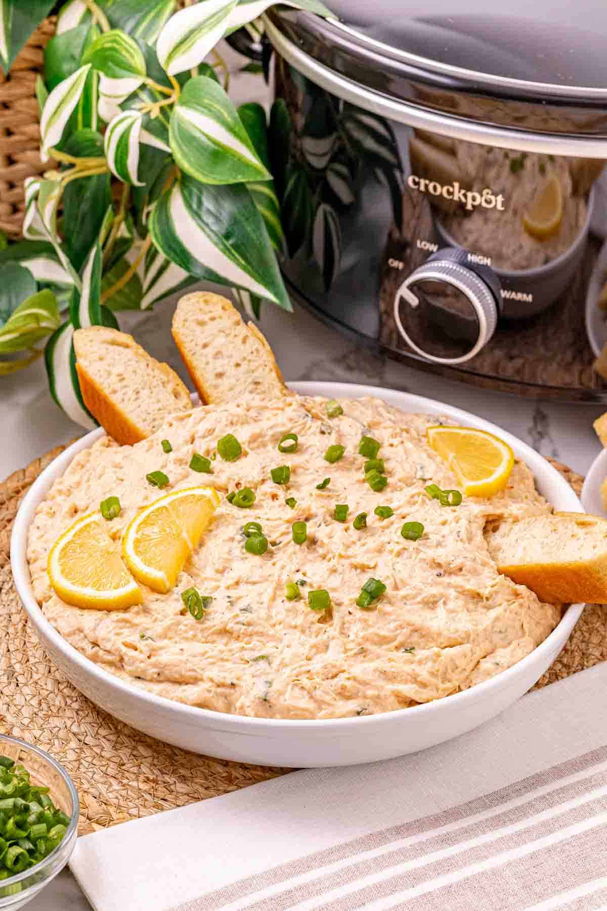 Creamy shredded chicken dip, reminiscent of a classic Crab Dip, garnished with lemon slices and green onions, served with bread slices near a Crockpot.