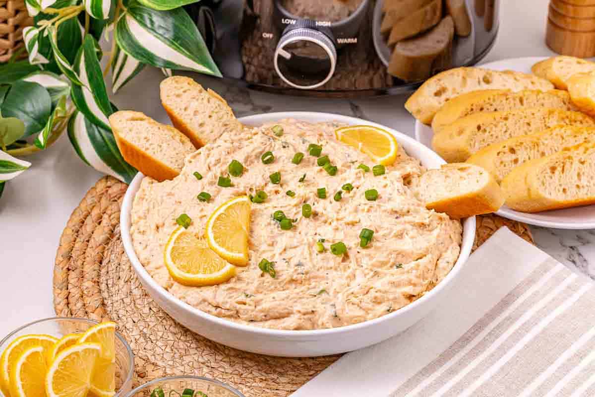A bowl of creamy crab dip recipe garnished with lemon slices, green onions, and baguette slices, surrounded by bread.