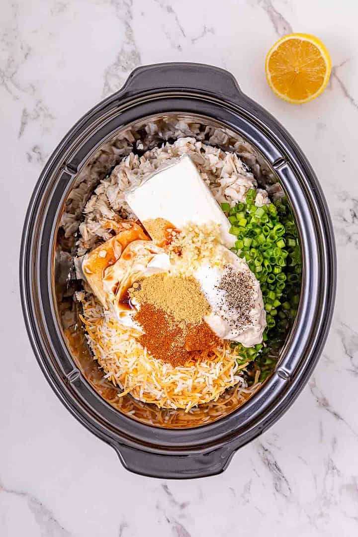 Creamy Crockpot dish featuring shredded chicken, cheese, spices, green onions, and cream cheese. A lemon half sits nearby to brighten the recipe with fresh flavor.
