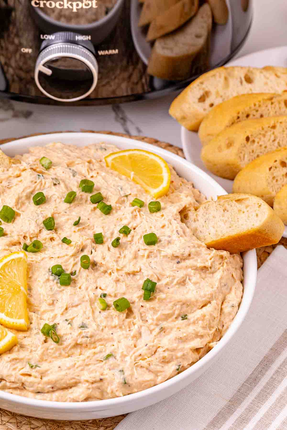 Creamy crab dip garnished with lemon slices and green onions, served with sliced baguette and a crockpot in the background&mdash;perfect for any gathering.