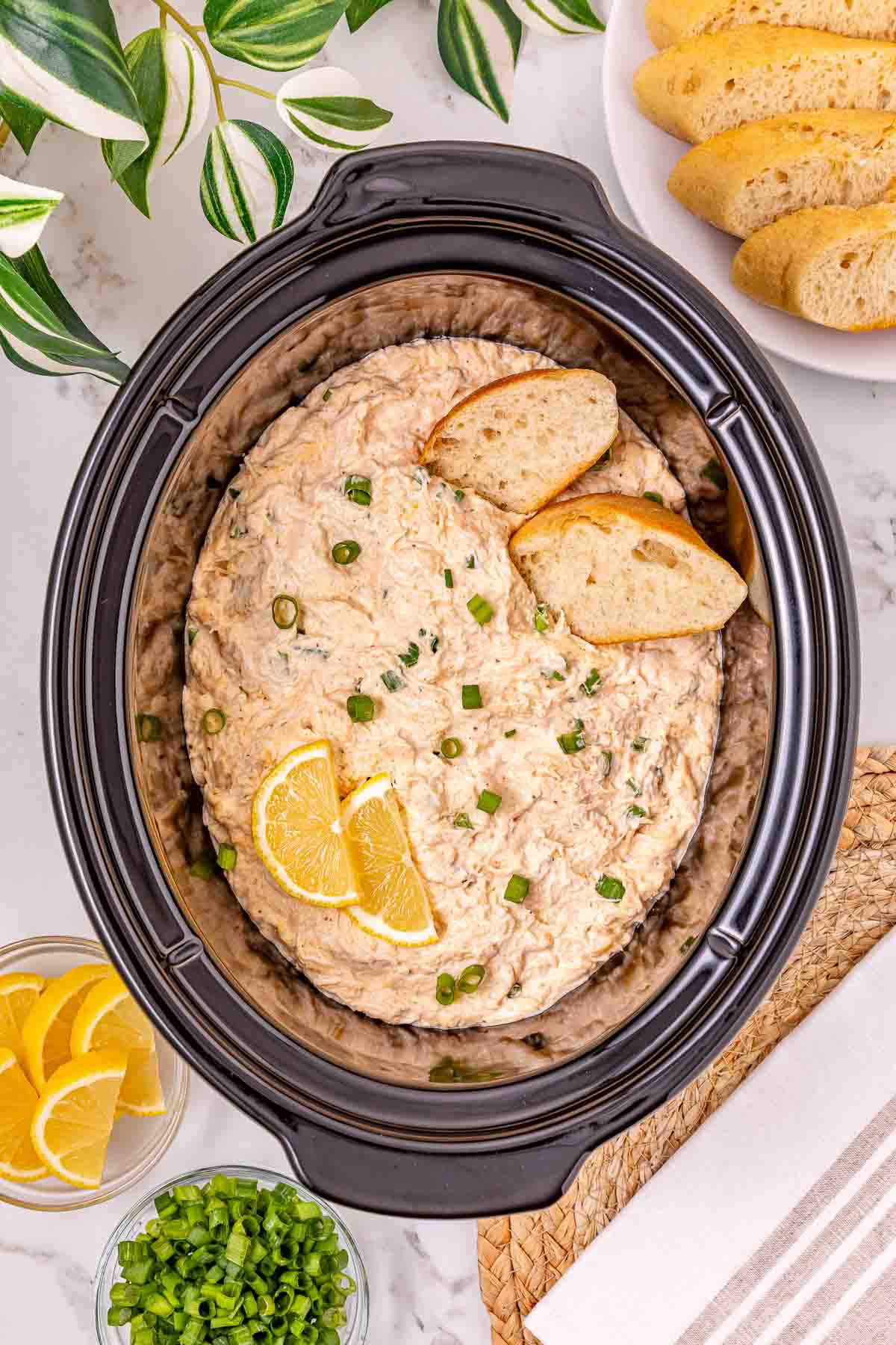 Crockpot of creamy crab dip topped with lemon slices, green onions, and bread slices, surrounded by bread and lemon wedges&mdash;a perfect recipe for sharing.
