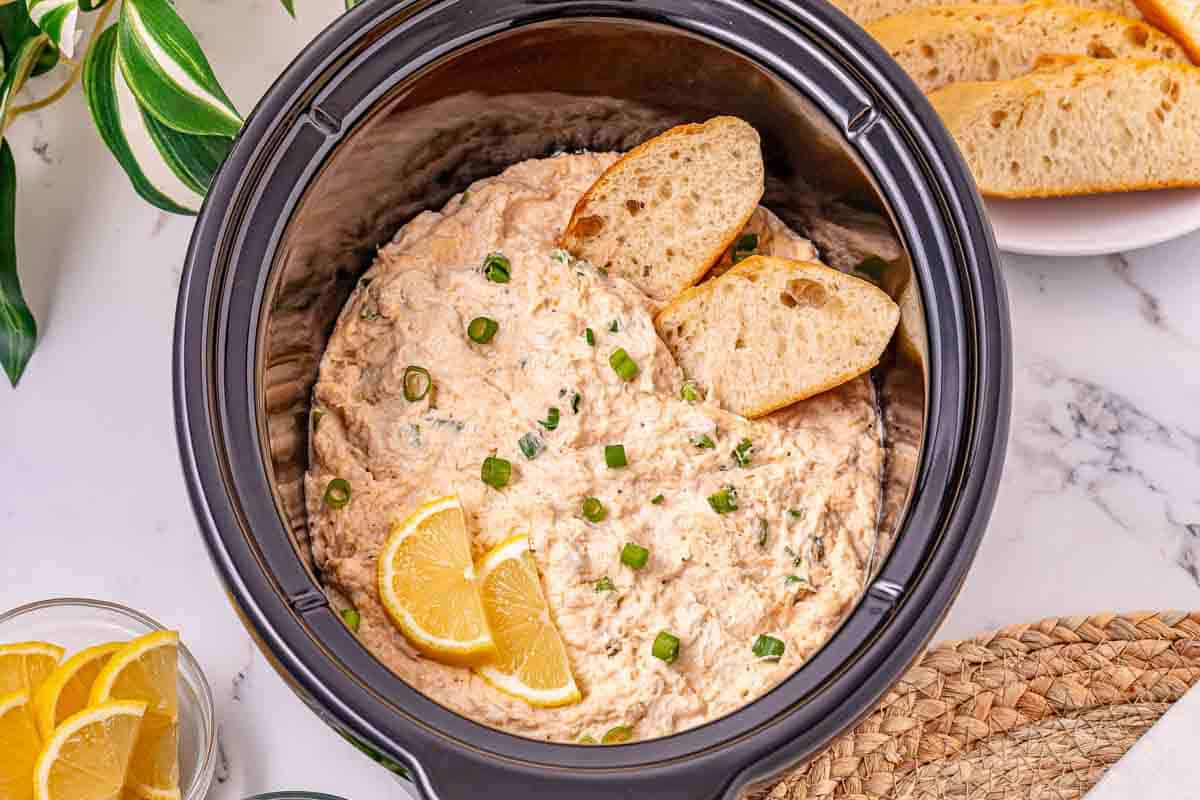 Creamy crab dip topped with green onions and lemon slices in a crockpot, served with bread slices for dipping&mdash;a must-try recipe for your next gathering.