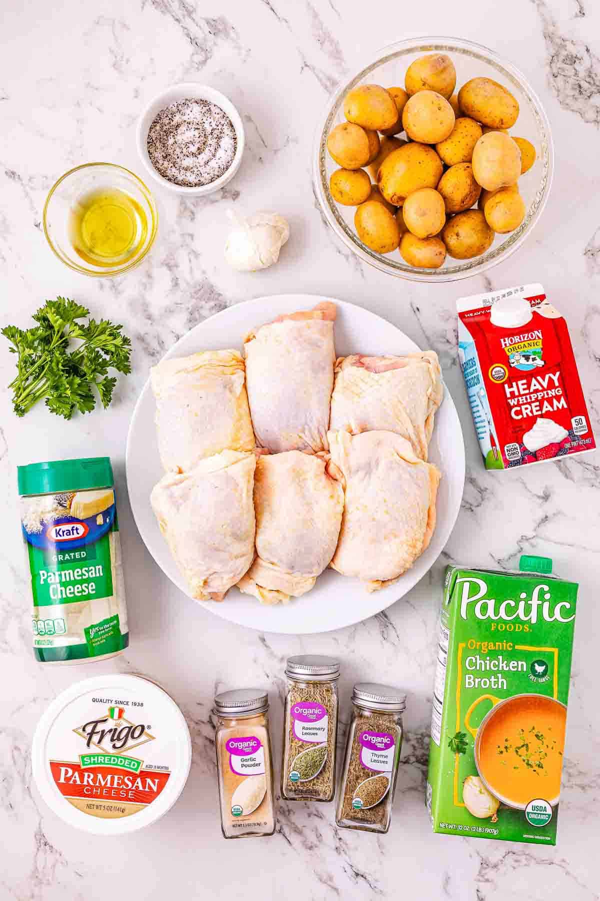 Raw chicken thighs, potatoes, cream, broth, parmesan, spices, oil, garlic, and parsley on a marble surface&mdash;perfect ingredients for savory slow cooker chicken or classic garlic parmesan chicken.