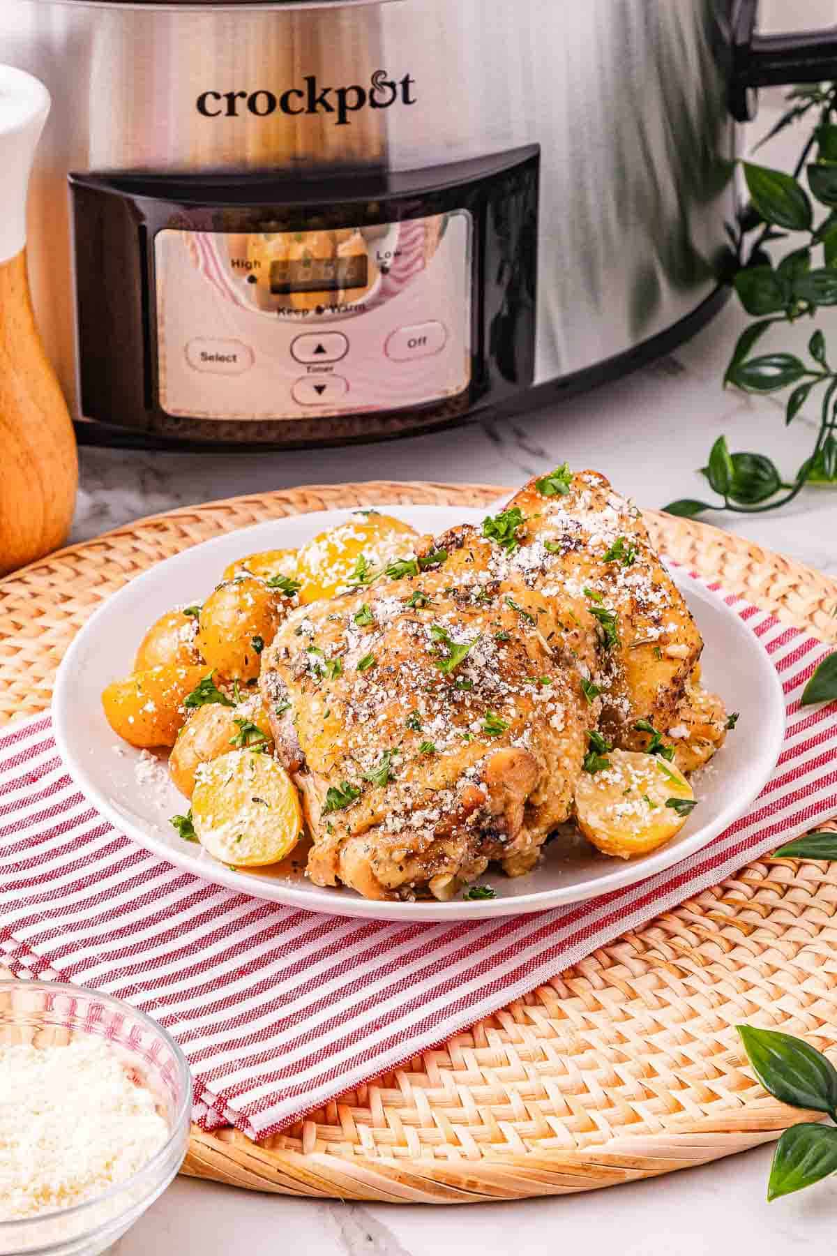 Plate of flavorful Crockpot Chicken and potatoes garnished with parsley, served in front of a crockpot on a striped napkin.