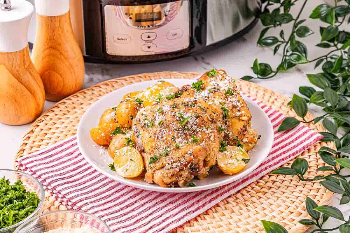 Plate of Garlic Parmesan chicken and potatoes garnished with herbs, with a slow cooker and greenery in the background.