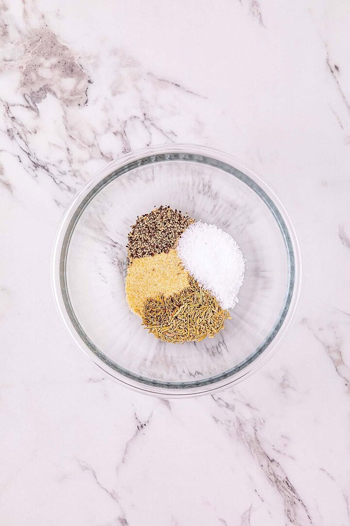 A glass bowl with dried herbs, black pepper, salt, and granulated garlic on a marble surface&mdash;perfect for seasoning Crockpot chicken or adding flavor to slow cooker chicken recipes.