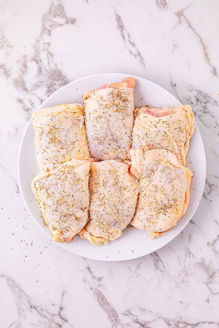 Six raw chicken thighs with skin, sprinkled with seasoning and ready for your favorite slow cooker chicken or garlic parmesan chicken recipe, arranged on a white plate on a marble surface.