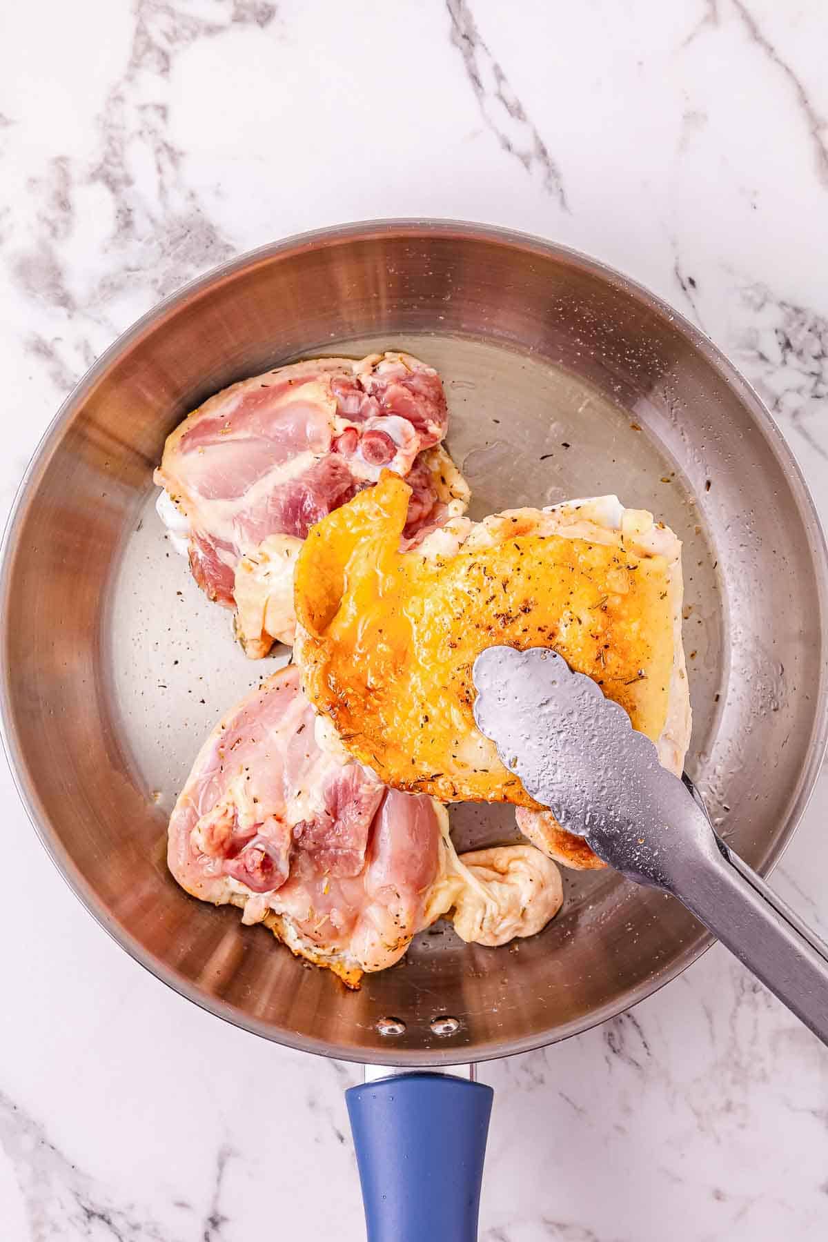Three raw chicken thighs are being seared in a stainless steel pan with tongs on a marble countertop, the perfect start for flavorful garlic parmesan chicken before finishing it in the Crockpot.