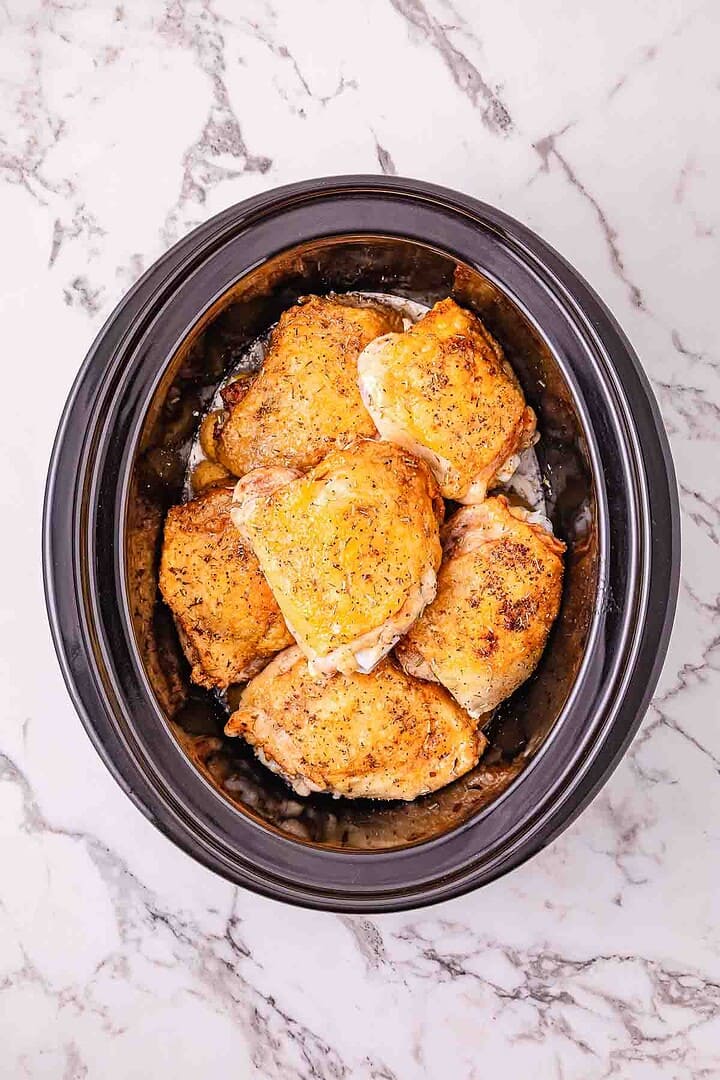 Six cooked garlic parmesan chicken thighs seasoned and arranged in a black Crockpot on a marble countertop.