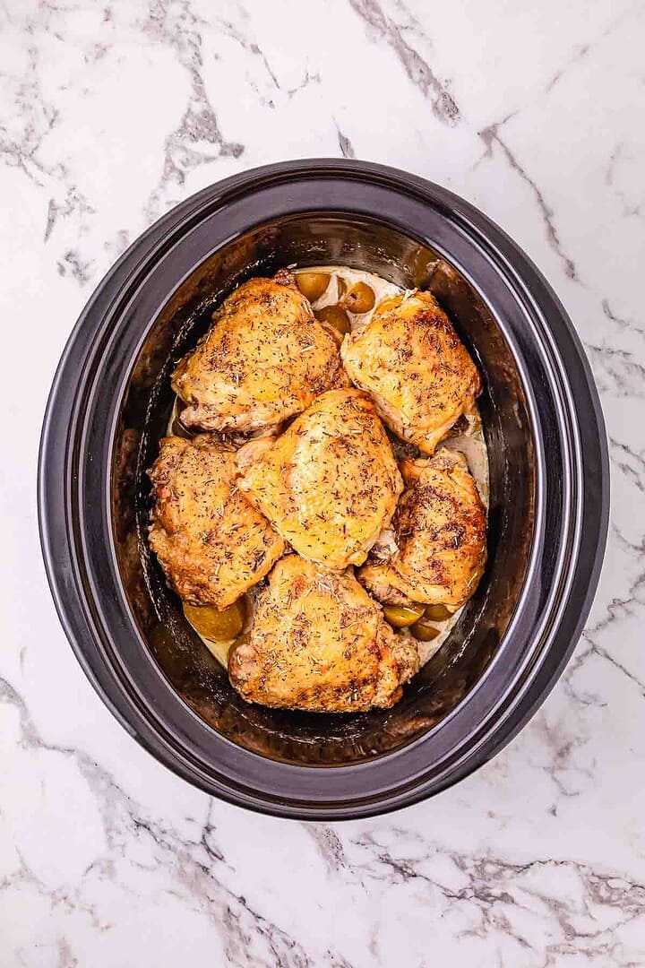 Six seasoned chicken thighs cooked in a black Crockpot on a marble countertop create the perfect slow cooker chicken dinner.