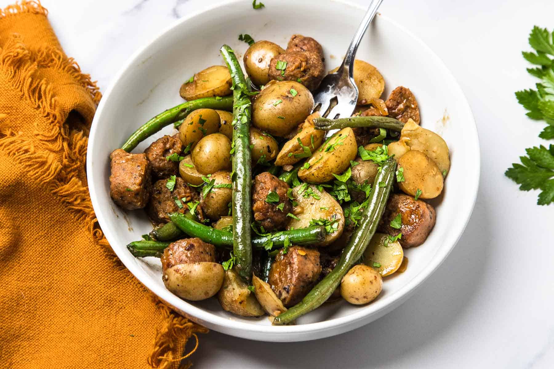 A bowl of Vegetarian Swamp Potatoes features green beans and baby potatoes garnished with herbs, with a fork and an orange napkin nearby.