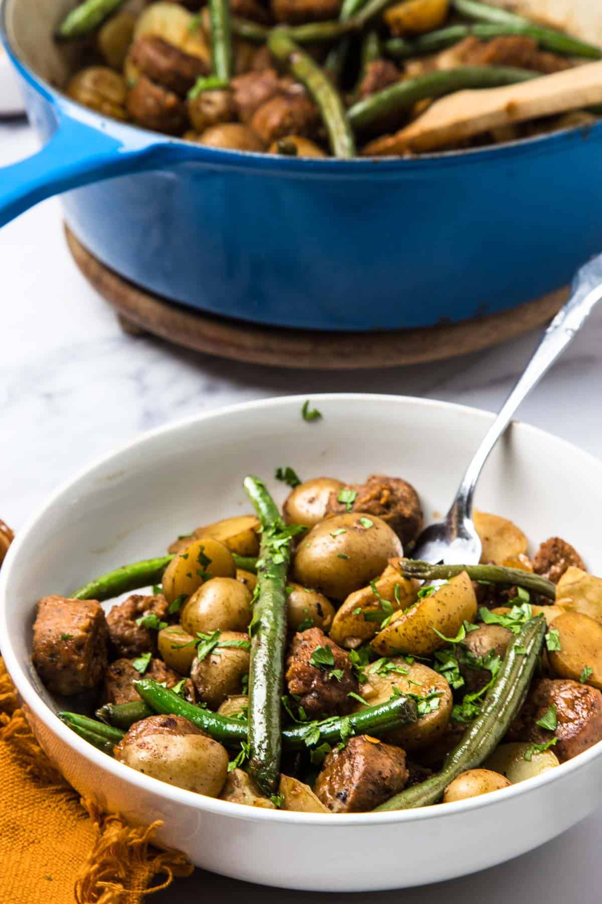 A white bowl of Vegetarian Swamp Potatoes&mdash;potatoes, green beans, and sausage&mdash;with a fork, in front of a blue pot filled with the same hearty dish.