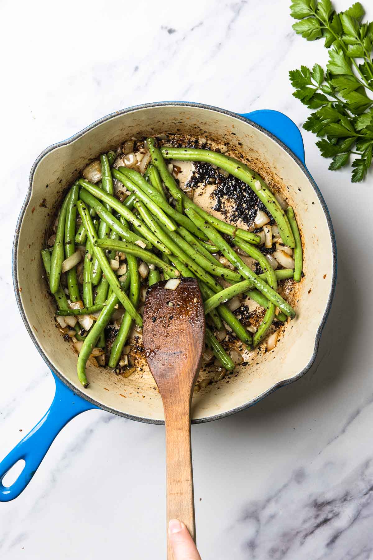 Green beans and chopped onions saut&eacute;ing in a blue skillet with a wooden spatula, perfect for pairing with Vegetarian Swamp Potatoes; fresh parsley on the side.