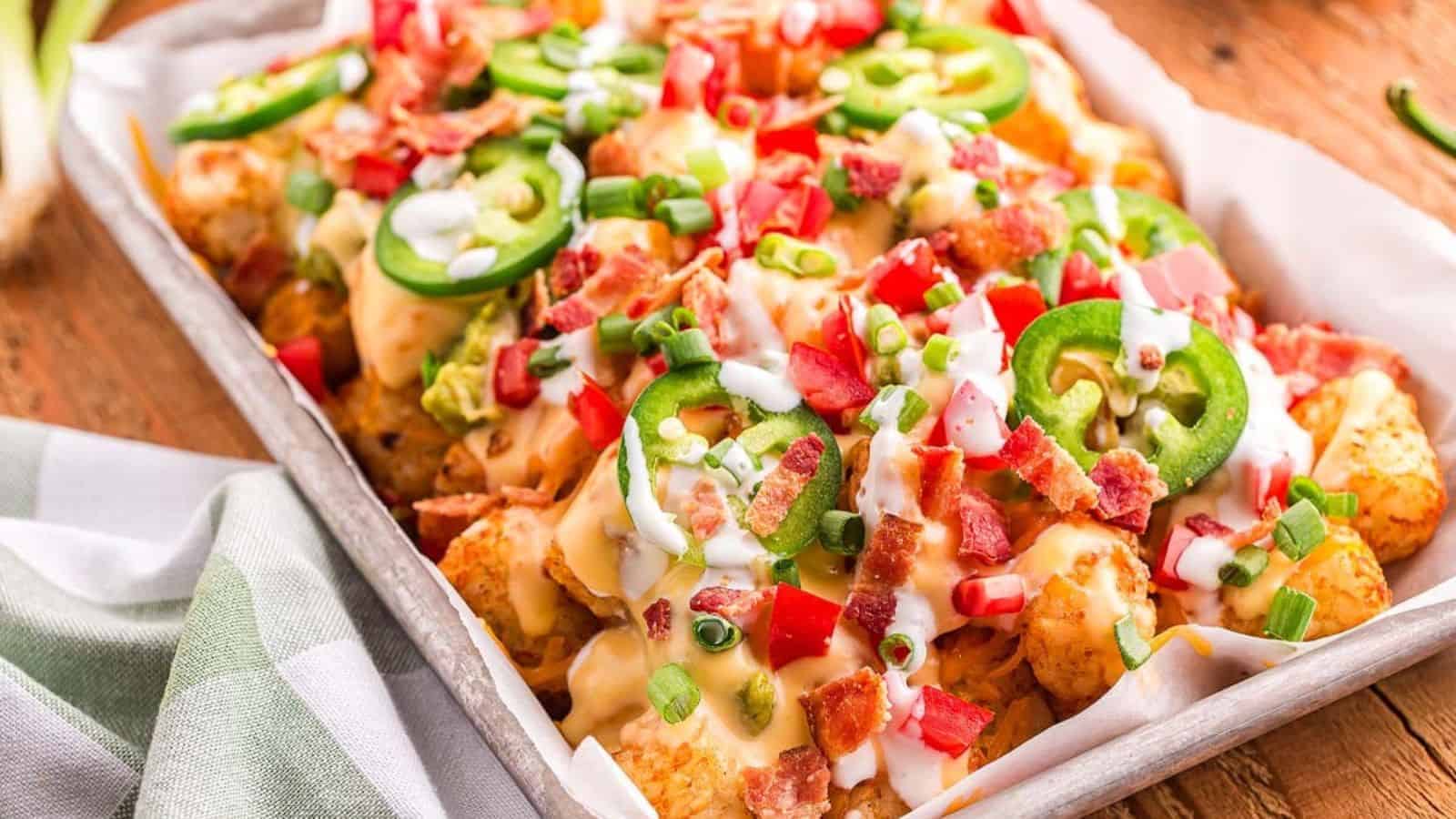 A tray of loaded tater tots topped with diced tomatoes, sliced jalape&ntilde;os, chopped green onions, bacon bits, and drizzled with melted cheese and sauce.