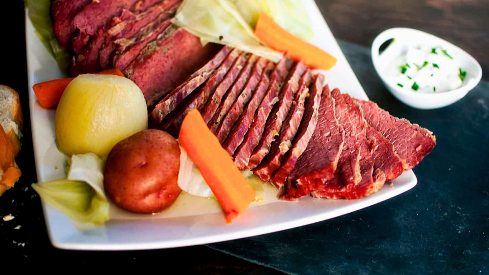 Sliced corned beef with potatoes, carrots, cabbage, and onions on a white platter, with sauce on the side.