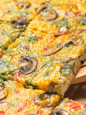 A wooden spatula lifts a slice of baked vegetable frittata cut into squares, topped with mushrooms and herbs.