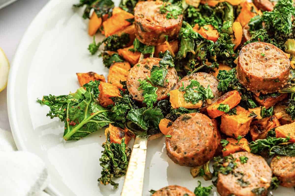 Sliced sausage, tender kale, and roasted sweet potatoes are served on a white plate, garnished with fresh herbs.