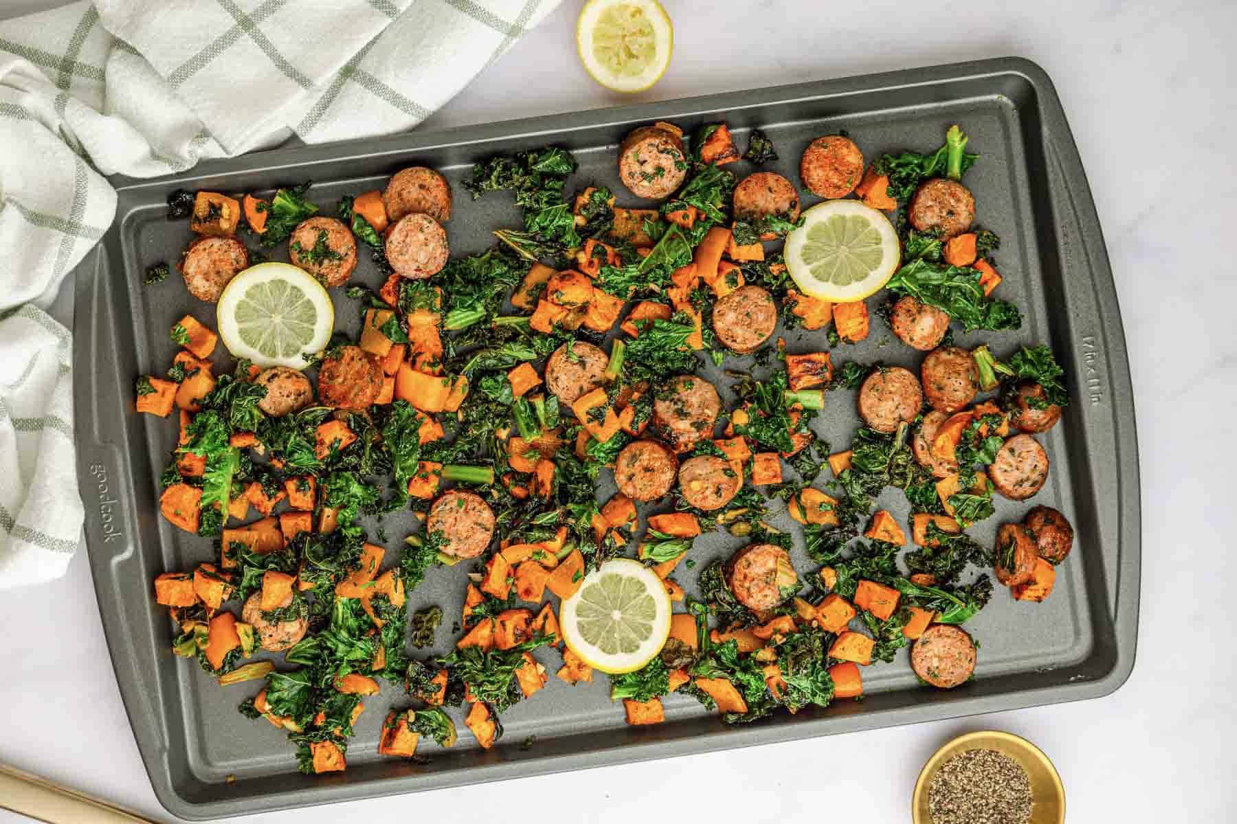 Sheet pan sausage with sliced kale, sweet potatoes, and lemon slices on a white background with a towel.
