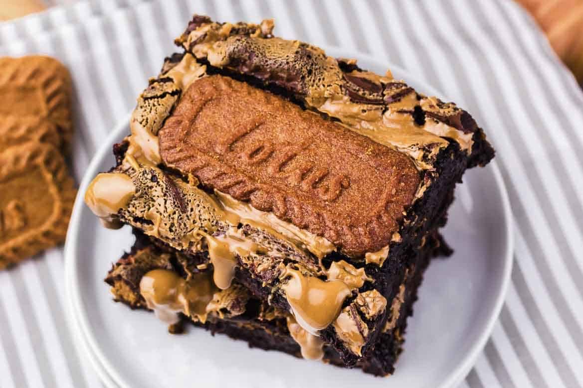 Two Biscoff brownies topped with Lotus biscuits and drizzled with caramel sauce on a white plate.