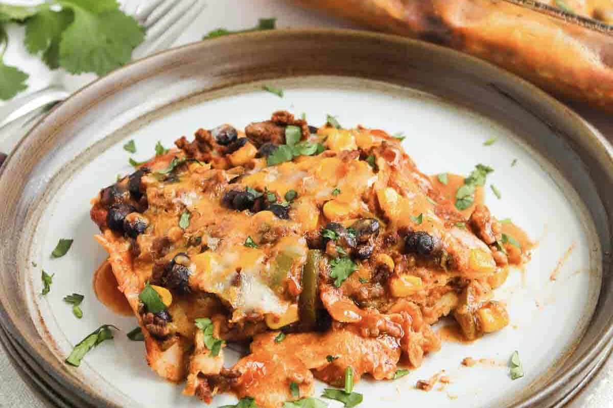 A plate of cheesy beef enchilada casserole with black beans, corn, and cilantro, served on a brown-rimmed dish&mdash;a classic beef enchilada casserole recipe.