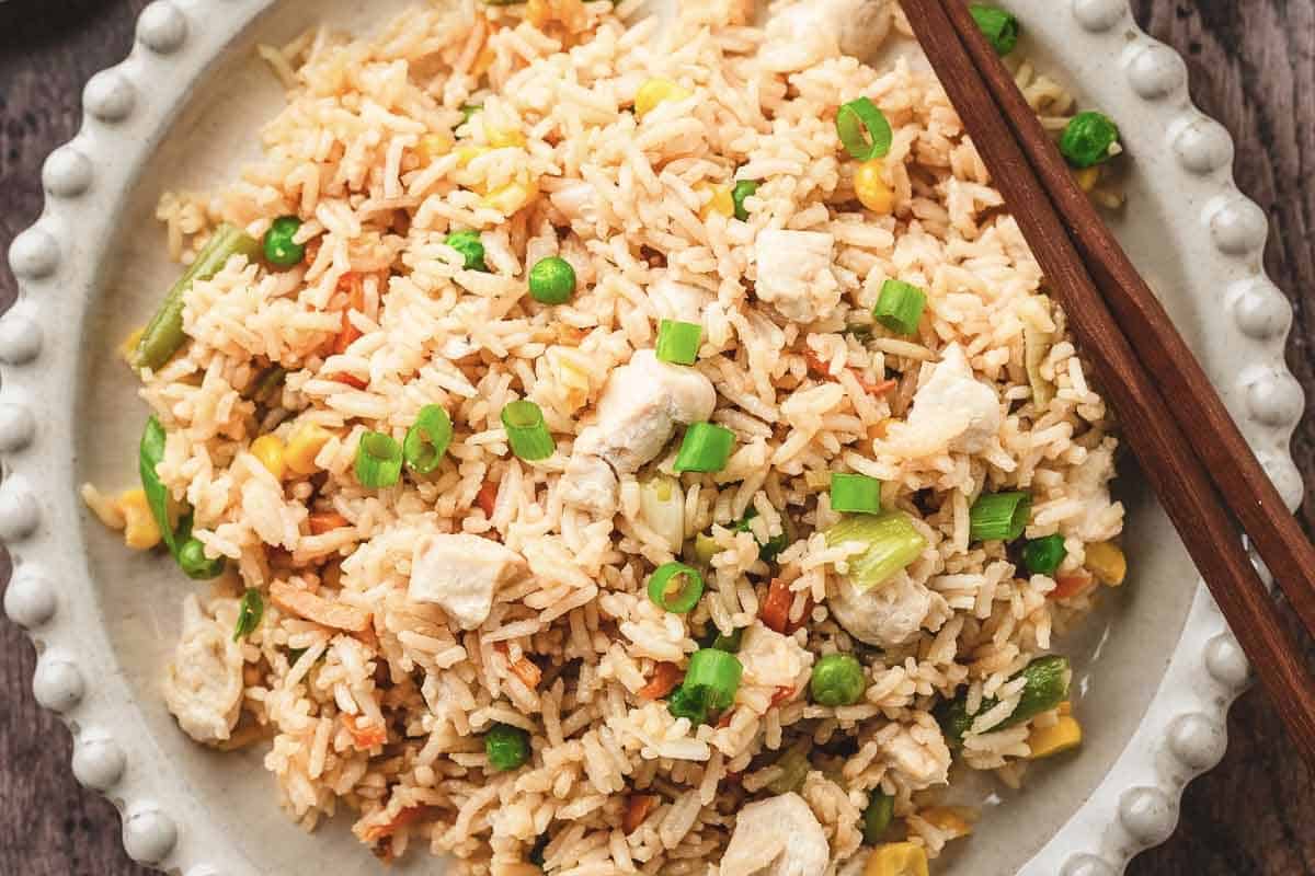 A plate of fried rice with vegetables, chicken pieces, and green onions, featuring a hint of teriyaki chicken flavor, served with chopsticks on the side.