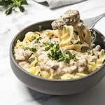 Bowl of creamy beef stroganoff with egg noodles, garnished with chopped parsley, on a white surface.