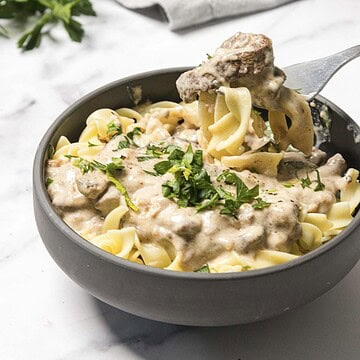 Bowl of creamy beef stroganoff with egg noodles, garnished with chopped parsley, on a white surface.