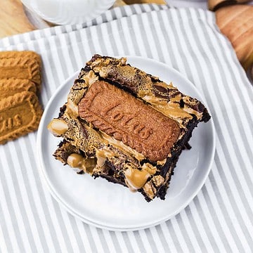 A chocolate brownie topped with a Lotus Biscoff cookie and caramel sauce on a white plate.