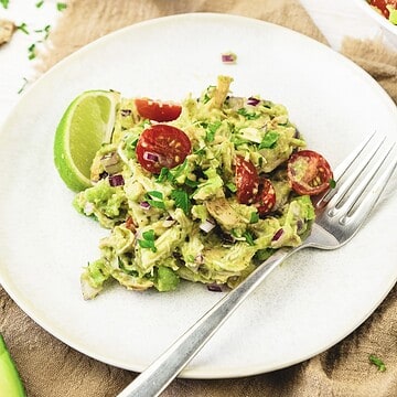 This chicken salad recipe features avocado, cherry tomatoes, red onion, and herbs, served with a lime wedge on a white plate.