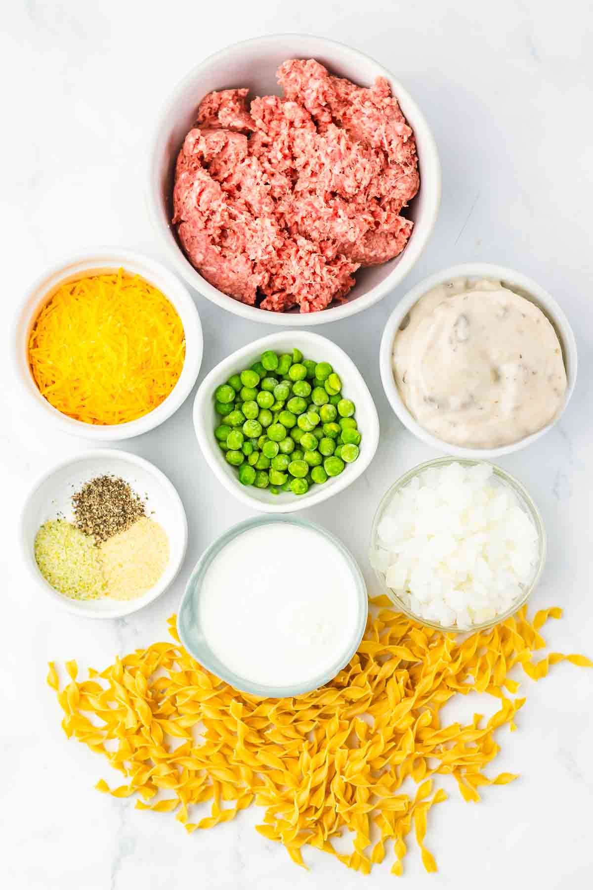 Bowls of ground beef, shredded cheese, peas, cream soup, spices, milk, chopped onion, and uncooked egg noodles&mdash;a hearty start for making classic Amish Country Casserole.