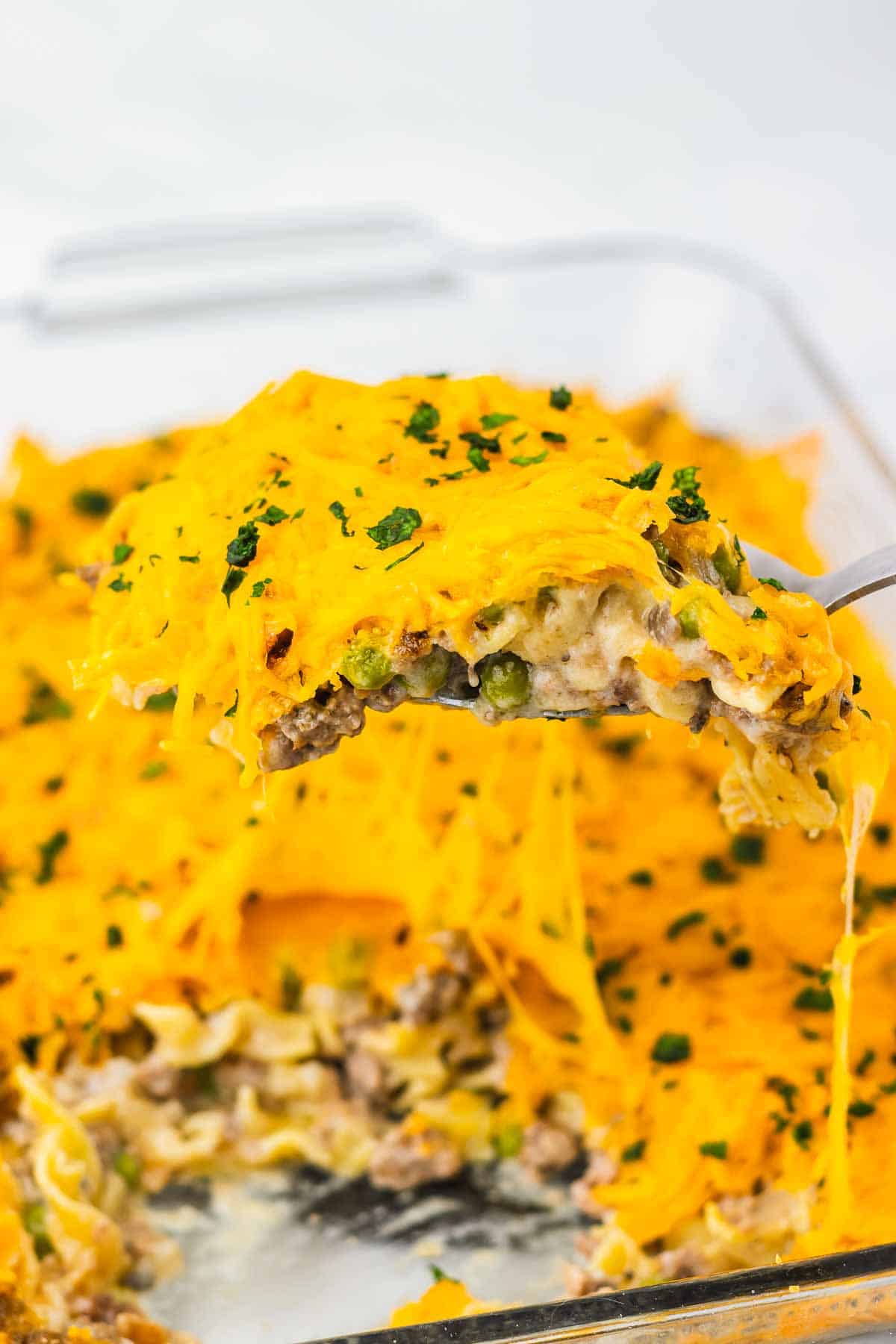A spatula lifts a cheesy Amish Country Casserole with beef, noodles, peas, and melted cheddar from a glass baking dish.