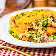 A plate of Amish Country Casserole with ground meat, peas, melted cheese, and herbs on top, served on a red checkered cloth.