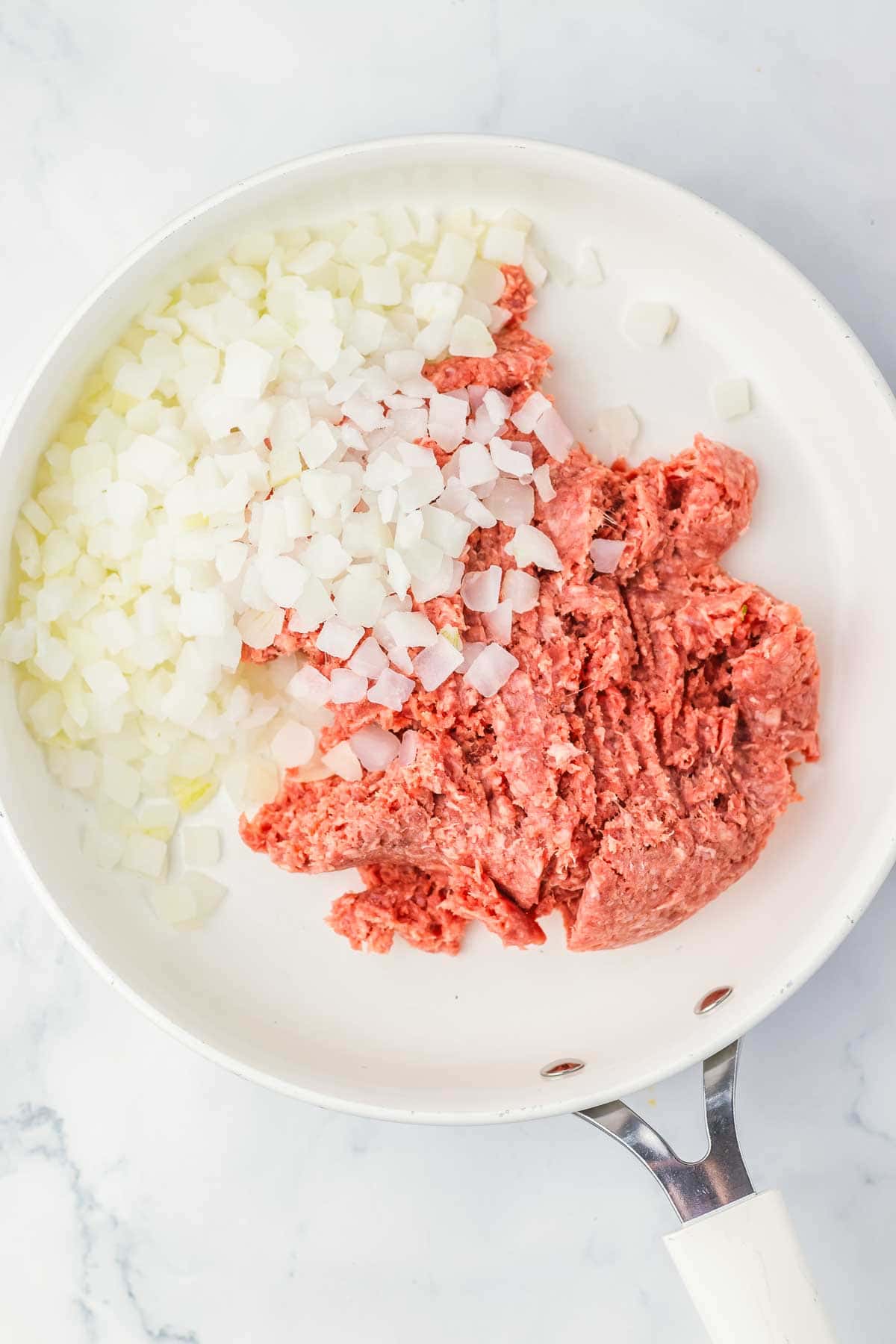 Raw ground beef and chopped onions in a white skillet on a marble countertop, ready to be cooked for a comforting Amish Country Casserole.