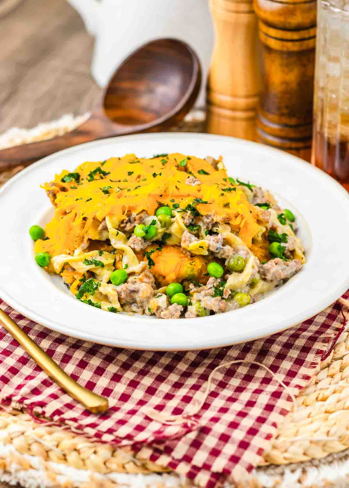 A plate of Amish Country Casserole topped with melted cheese, peas, and parsley, on a checkered napkin with a gold fork.