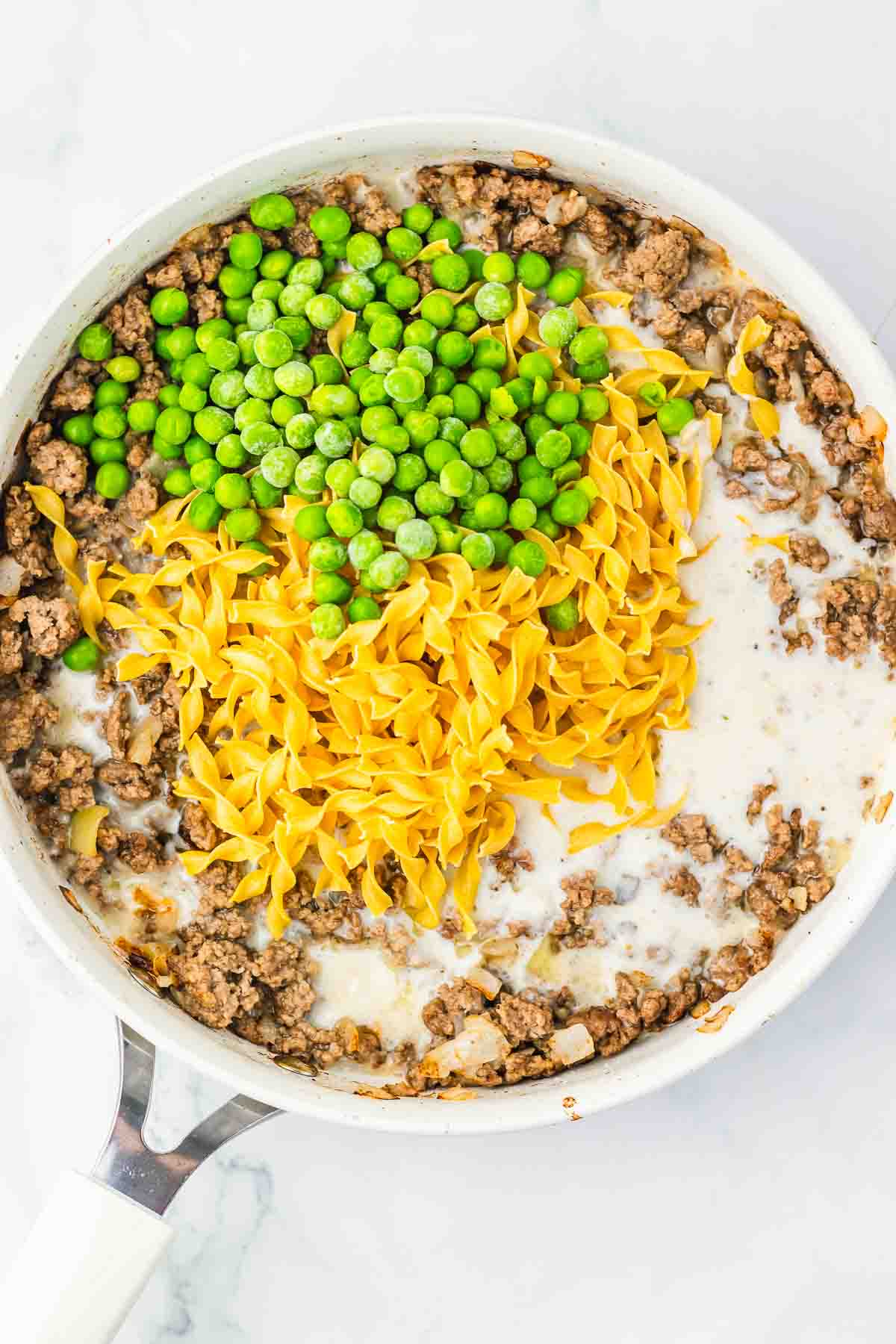A skillet with ground beef, egg noodles, green peas, and creamy sauce comes together for a comforting Amish Country Casserole on a white background.