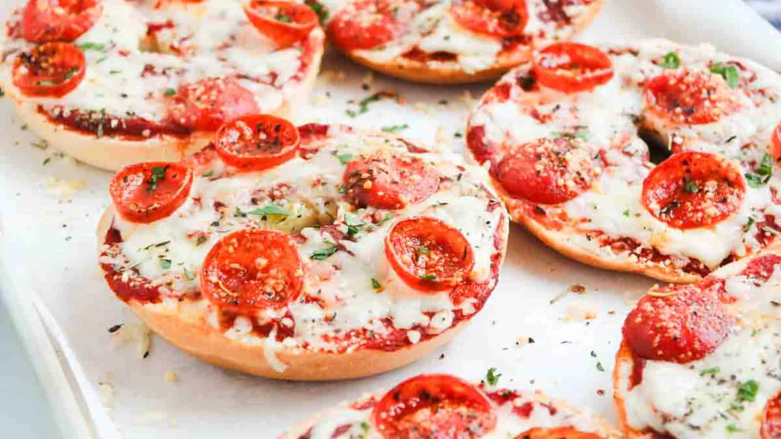 Bagels topped with melted cheese, tomato sauce, and pepperoni, garnished with herbs on a baking tray.