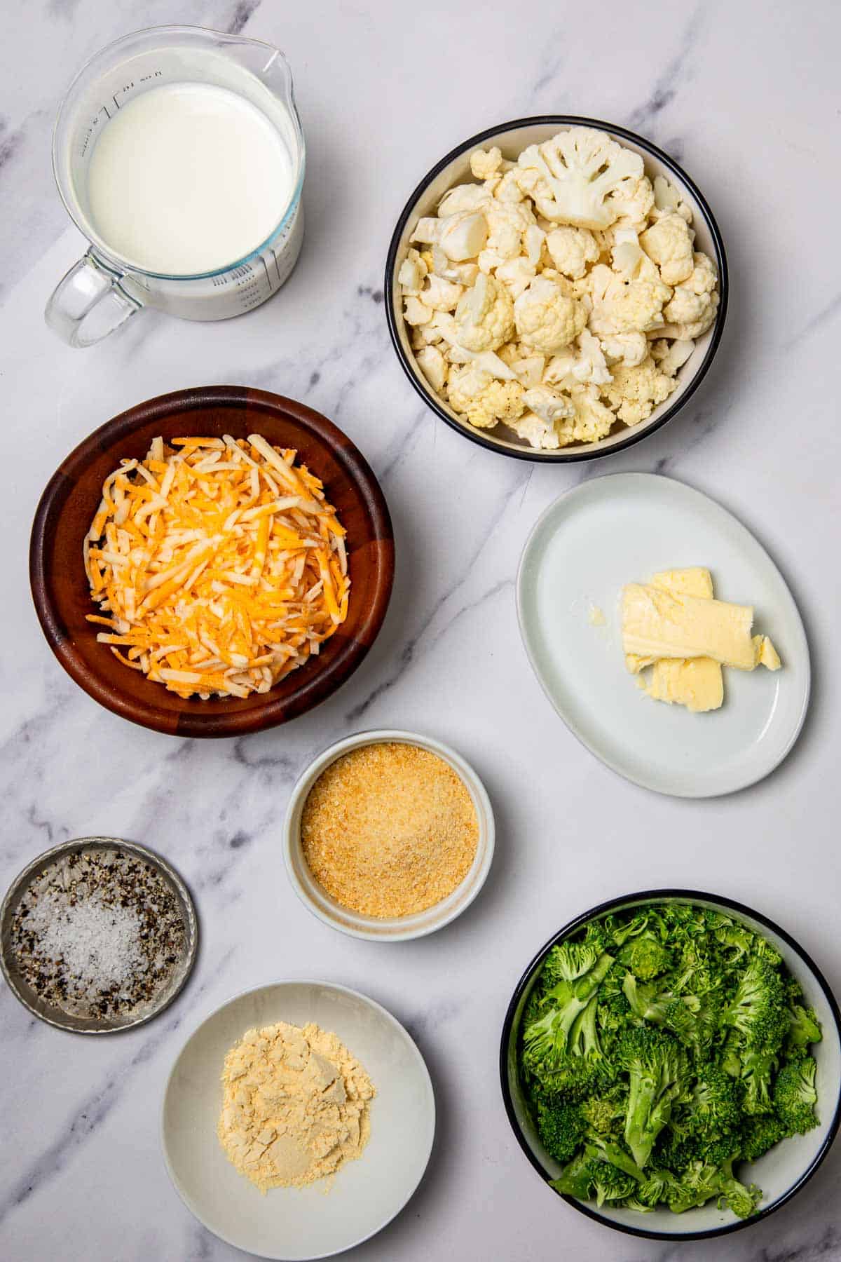 Milk, butter, shredded cheese, cauliflower, broccoli, bread crumbs, flour, salt, and pepper are ready on a marble surface for a delicious Cheese Casserole.