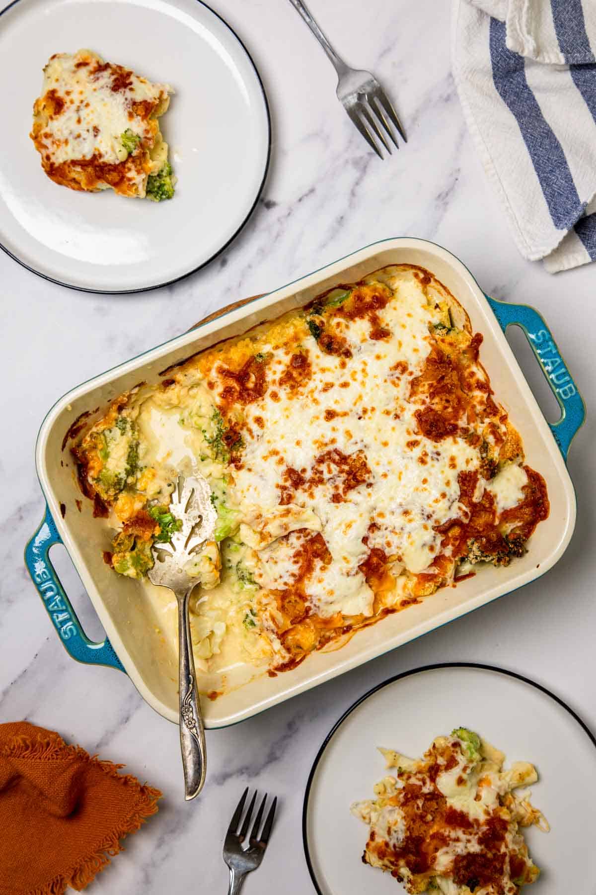 A cheesy baked Cheese Casserole loaded with broccoli and cauliflower, served in a dish with two portions plated on a marble table, utensils nearby.
