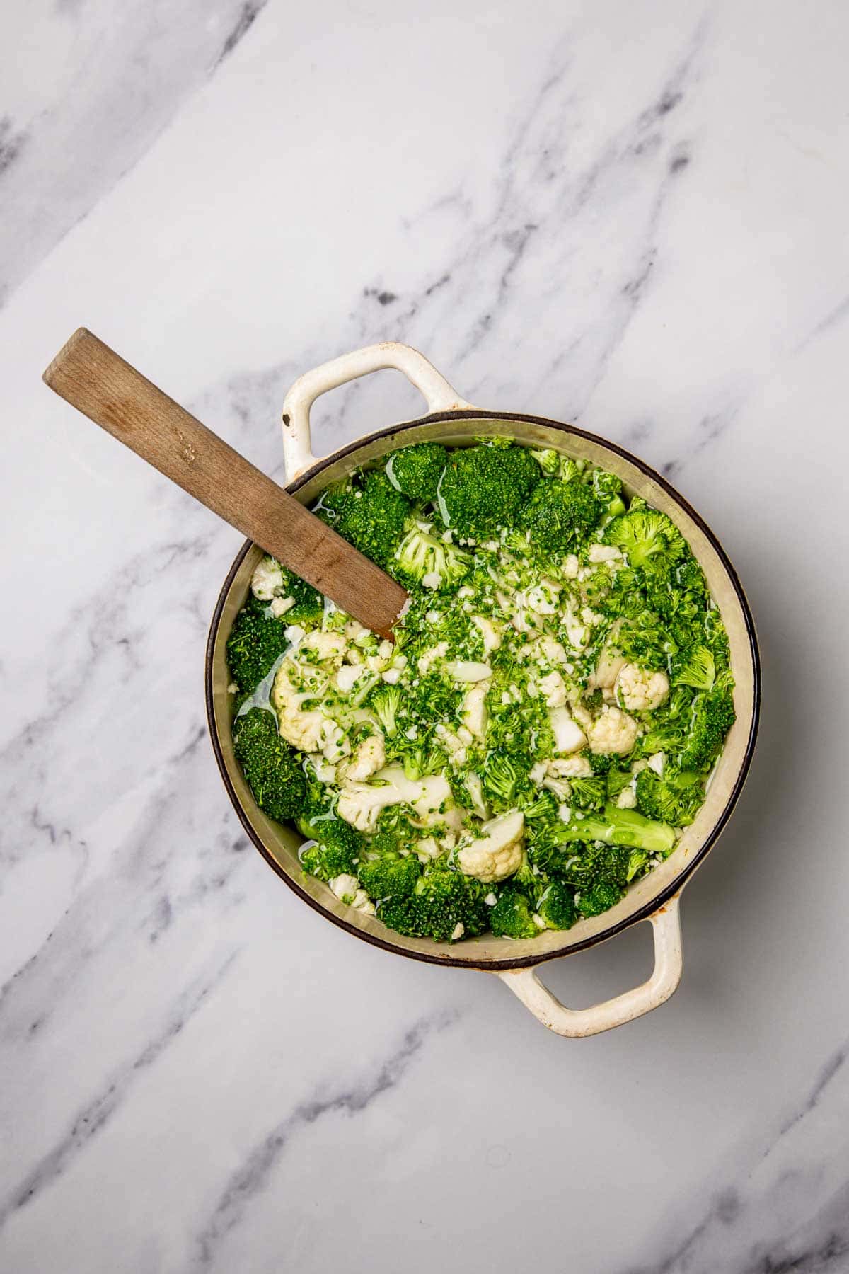 Large white pot with broccoli, cauliflower, and green sauce on a marble surface; perfect for making a cheesy broccoli and cauliflower casserole. Wooden spoon inside.