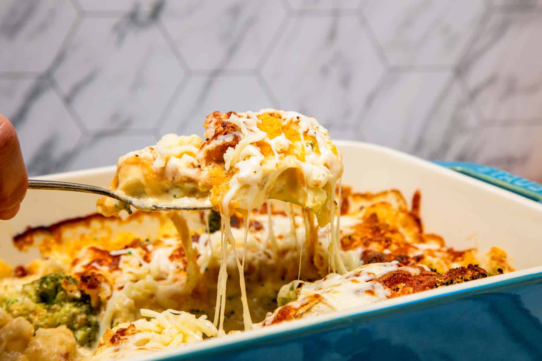 A spoon lifts cheesy, baked Cheese Casserole from a blue dish, with melted cheese stretching&mdash;filled with tender broccoli and cauliflower.