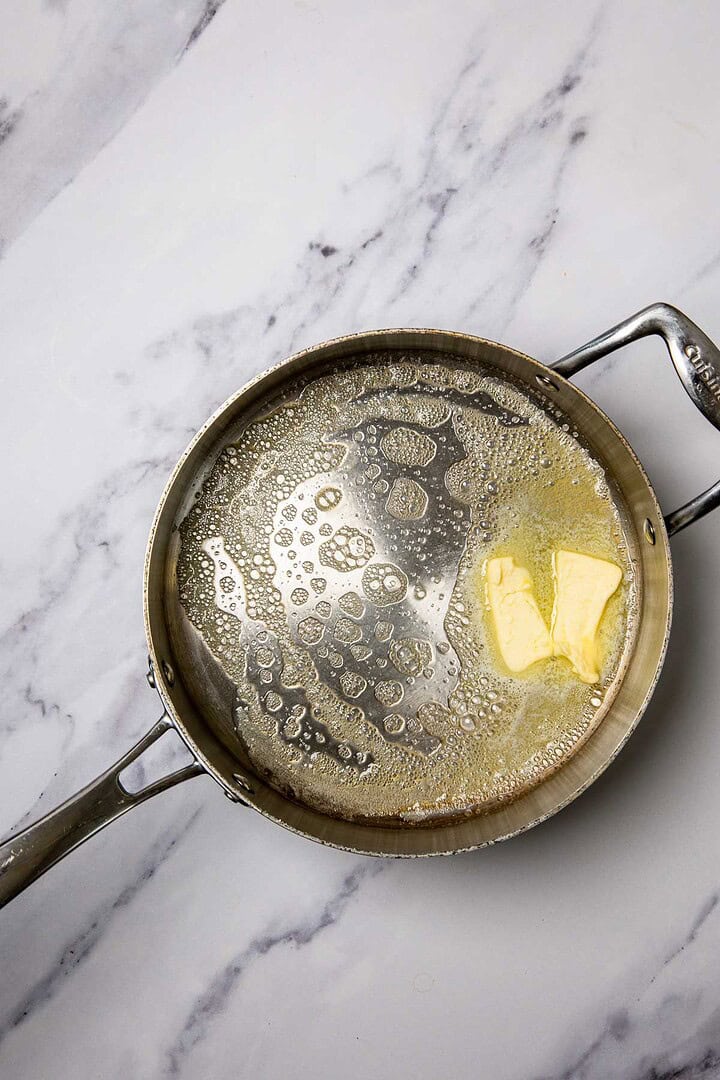 A metal skillet with melting butter on a marble countertop, perfect for starting a delicious cheese casserole with broccoli and cauliflower.