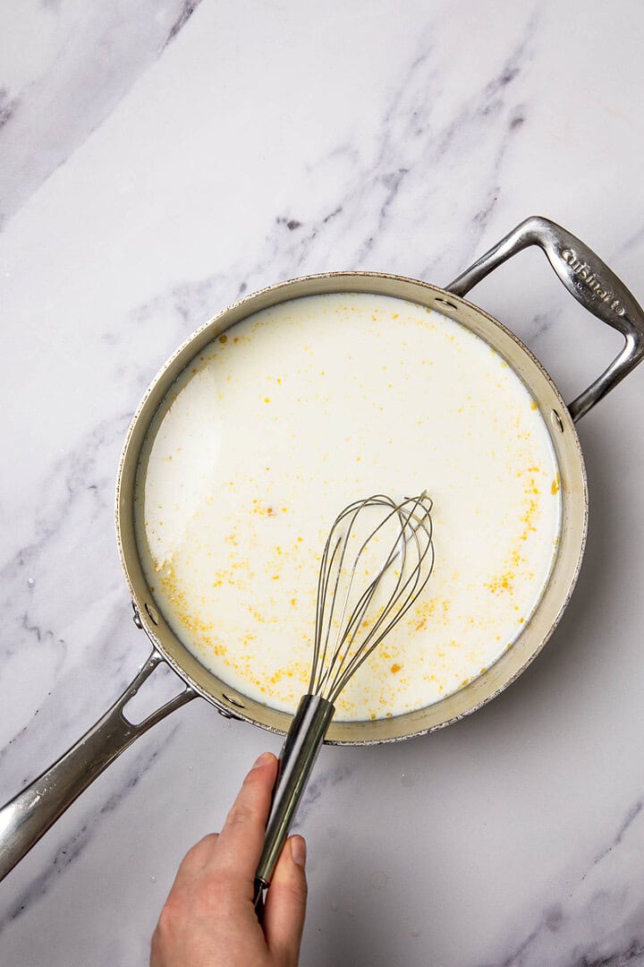 A hand whisking milk with yellow specks for a Cheese Casserole in a stainless steel pan on a marble countertop.