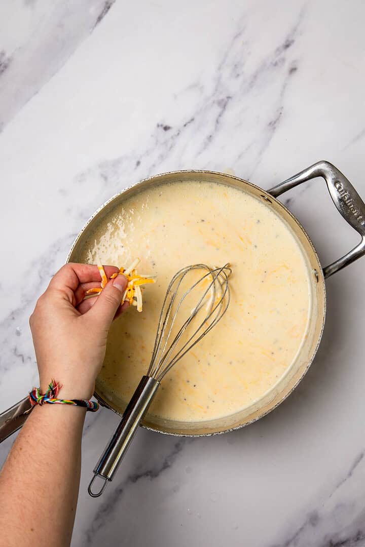 A hand adds shredded cheese to a creamy sauce in a pan with a whisk on a marble countertop, ready to create a cheesy Broccoli and Cauliflower Casserole.